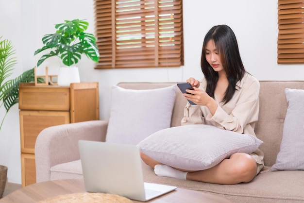 Koncepcja relaksu w domu Młoda Azjatka używa smartfona do rozmowy z przyjaciółmi na kanapie