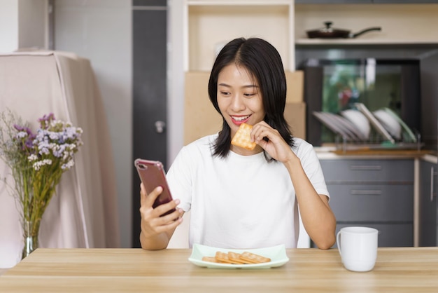 Koncepcja relaksu w domu Młoda kobieta je krakersy i surfuje w mediach społecznościowych na smartfonie w domu