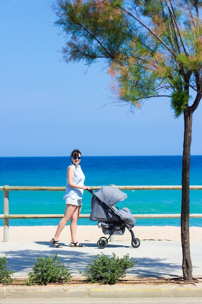 Koncepcja Rodzicielstwa I Podróży - Widok Z Boku Młodej Kobiety Pchającej Wózek Dziecięcy Na Letniej Plaży