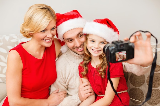 koncepcja rodziny, bożego narodzenia, bożego narodzenia, zimy, szczęścia i ludzi - uśmiechnięta rodzina w czapkach pomocnika mikołaja robi zdjęcie aparatem fotograficznym