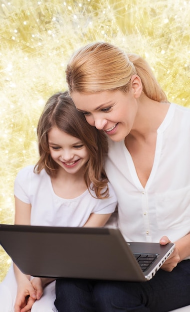 koncepcja rodziny, dzieciństwa, wakacji, technologii i ludzi - uśmiechnięta matka i mała dziewczynka z laptopem na żółtym tle światła