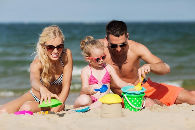 koncepcja rodziny, podróży, wakacji i ludzi - szczęśliwy mężczyzna, kobieta i dziewczynka w okularach przeciwsłonecznych bawiących się zabawkami z piasku na letniej plaży