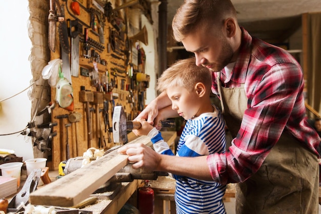 koncepcja rodziny, stolarstwa, rzemiosła i ludzi - ojciec i mały syn z młotkiem uderzają gwoździem w drewnianą deskę w warsztacie