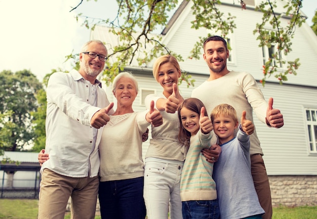 koncepcja rodziny, szczęścia, pokolenia, domu i ludzi - szczęśliwa rodzina stojąca przed domem i pokazująca kciuk w górę na zewnątrz