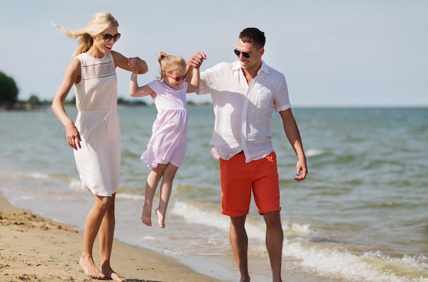 koncepcja rodziny, wakacji, adopcji i ludzi - szczęśliwy mężczyzna, kobieta i dziewczynka w okularach przeciwsłonecznych chodzących po letniej plaży