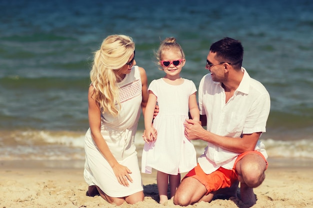 koncepcja rodziny, wakacji, adopcji i ludzi - szczęśliwy mężczyzna, kobieta i dziewczynka w okularach przeciwsłonecznych rozmawiających na letniej plaży