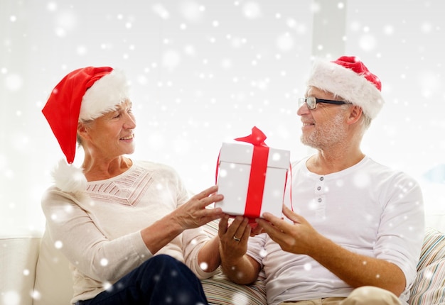 koncepcja rodziny, wakacji, świąt, wieku i ludzi - szczęśliwa para starszych w czapkach mikołaja z pudełkiem prezentów w domu