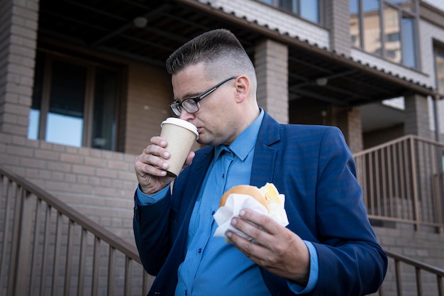 Koncepcja śmieciowego jedzenia na przerwie obiadowej Biznesmen zjada niezdrowe, wysokokaloryczne jedzenie na ulicy na tle budynku biurowego
