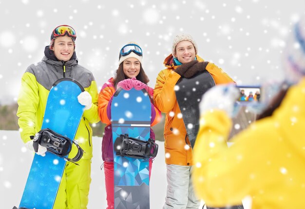 Zdjęcie koncepcja sportów zimowych, technologii, wypoczynku, przyjaźni i ludzi - szczęśliwi przyjaciele ze snowboardami i smartfonem robiący zdjęcia na zewnątrz