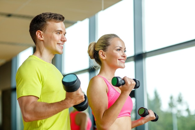 koncepcja sportu, fitnessu, stylu życia i ludzi - uśmiechnięty mężczyzna i kobieta z hantlami ćwiczącymi na siłowni