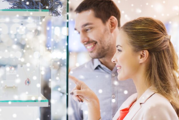 koncepcja sprzedaży, konsumpcjonizmu i ludzi - szczęśliwa para wskazująca palcem na okno sklepu jubilerskiego w centrum handlowym z efektem śniegu