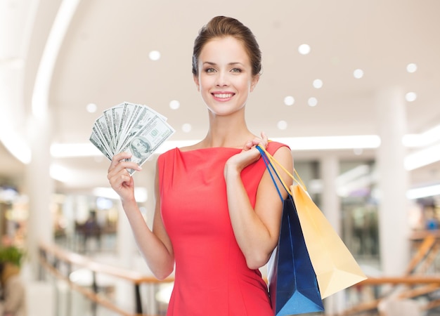 koncepcja sprzedaży, pieniędzy, ludzi i wakacji - uśmiechnięta kobieta w czerwonej sukience z torbami na zakupy i dolarami na tle centrum handlowego