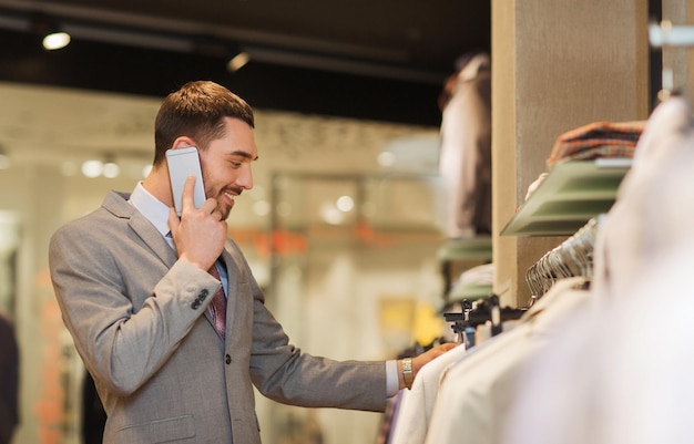 koncepcja sprzedaży, zakupów, mody, komunikacji i ludzi - szczęśliwy młody człowiek dzwoni na smartfona i wybiera ubrania w sklepie odzieżowym