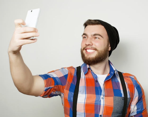 Koncepcja stylu życia: młody człowiek z brodą w koszuli, trzymając telefon komórkowy i robiąc sobie zdjęcie, stojąc na szarym tle.