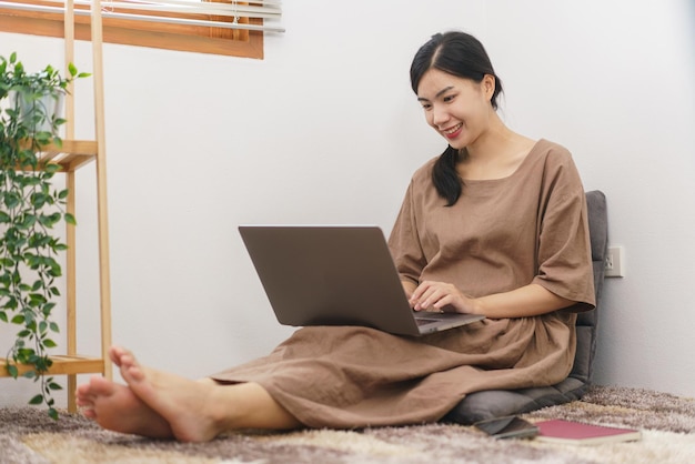 Koncepcja stylu życia relaksacyjnego Młoda Azjatycka kobieta używa laptopa do surfowania po mediach społecznościowych w salonie