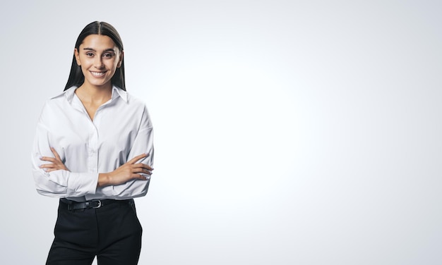 Koncepcja sukcesu w biznesie z młoda uśmiechnięta kobieta składa ręce w białej koszuli i czarnych spodniach na jasnym pustym tle z miejscem na logo lub tekst makiety