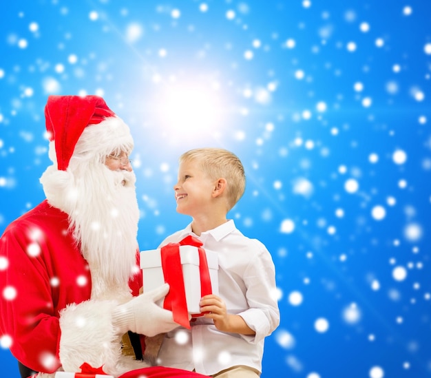 koncepcja świąt, bożego narodzenia, dzieciństwa i ludzi - uśmiechnięty mały chłopiec ze świętym mikołajem i prezentami na niebieskim śnieżnym tle