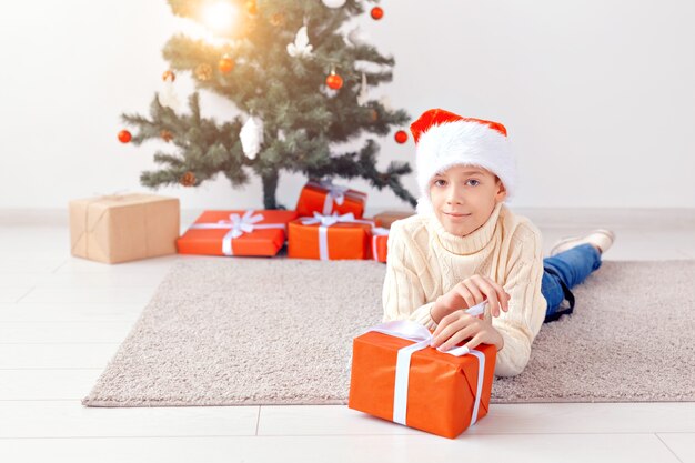 Koncepcja święta, boże narodzenie, dzieciństwo i ludzie - uśmiechnięty szczęśliwy chłopiec teen w santa hat otwiera pudełko na tle choinki.