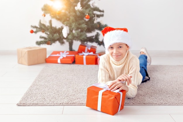 Koncepcja święta, boże narodzenie, dzieciństwo i ludzie - uśmiechnięty szczęśliwy chłopiec teen w santa hat otwiera pudełko na tle choinki.