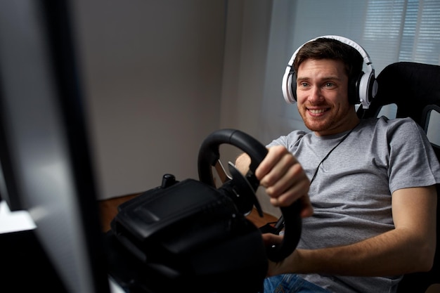 koncepcja technologii, gier, rozrywki i ludzi - szczęśliwy uśmiechnięty młody człowiek w słuchawkach z komputerem PC grający w wyścigi samochodowe w domu i na kierownicy