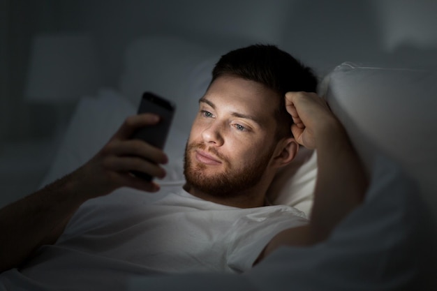 koncepcja technologii, internetu, komunikacji i ludzi - młody człowiek pisze SMS-y na smartfonie w łóżku w domu w nocy