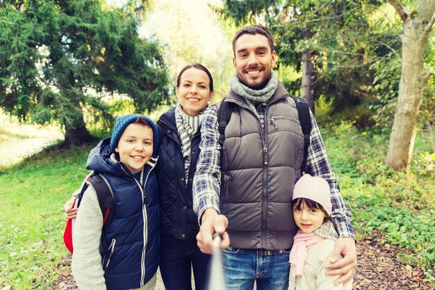 koncepcja technologii, podróży, turystyki, wędrówek i ludzi - szczęśliwa rodzina z plecakami robiąca zdjęcia za pomocą selfie stick i pieszych wędrówek