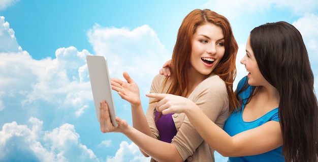 koncepcja technologii, przyjaźni i ludzi - dwie uśmiechnięte nastolatki lub młode kobiety wskazujące palcem na komputer typu tablet PC nad błękitnym niebem z chmurami w tle