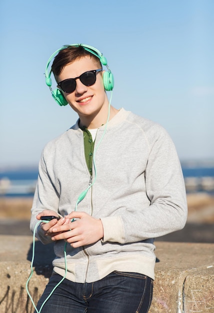 koncepcja technologii, stylu życia i ludzi - uśmiechnięty młody mężczyzna lub nastoletni chłopak w słuchawkach ze smartfonem słuchający muzyki na świeżym powietrzu