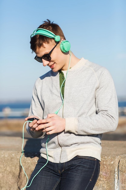 koncepcja technologii, stylu życia i ludzi - uśmiechnięty młody mężczyzna lub nastoletni chłopak w słuchawkach ze smartfonem słuchający muzyki na świeżym powietrzu