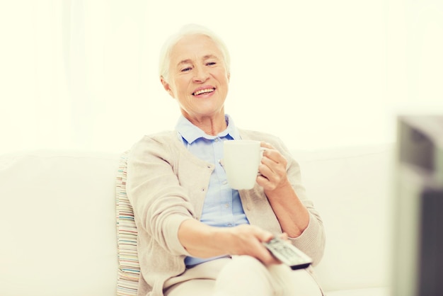 koncepcja technologii, telewizji, wieku i ludzi - szczęśliwa starsza kobieta ogląda telewizję, pije herbatę i zmienia kanały za pomocą pilota w domu