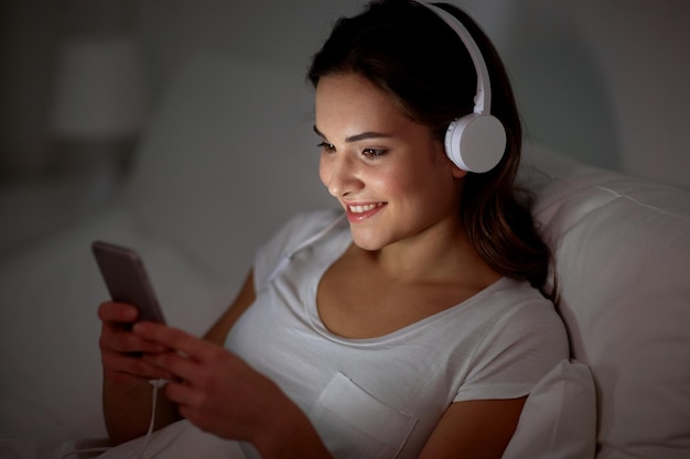 Zdjęcie koncepcja technologii, wypoczynku i ludzi - szczęśliwa uśmiechnięta młoda kobieta ze smartfonem i słuchawkami słuchająca muzyki w łóżku w nocy