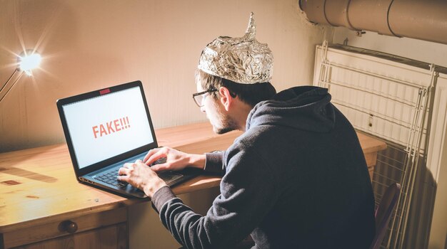 Koncepcja teorii spiskowej młody człowiek z aluminiową czapką przeszukujący internet siedzący samotnie w ciemnej piwnicy