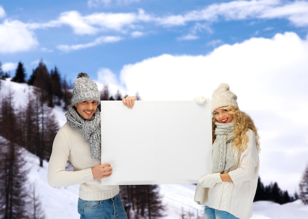 Koncepcja Turystyki, Ferii Zimowych, świąt, Reklamy I Ludzi - Uśmiechnięta Para W Zimowych Ubraniach Trzymająca Dużą Białą Pustą Deskę Nad Tłem Zaśnieżonych Gór