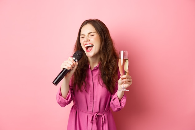 Koncepcja uroczystości i wakacji. Wizerunek imprezowiczki, która dobrze się bawi, pije szampana i śpiewa karaoke, trzyma mikrofon, stoi szczęśliwa przed różową ścianą.