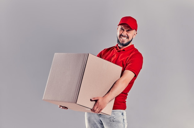 Koncepcja usługi kurierskiej i kurierskiej. Przystojny facet skrzywiony stara się trzymać w ręku duże kartonowe pudełko, na białym tle na szarym tle.