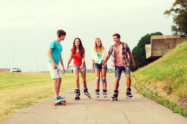 koncepcja wakacje, wakacje, miłość i przyjaźń - grupa uśmiechniętych nastolatków z wrotkami i deskorolką jeżdżących na świeżym powietrzu