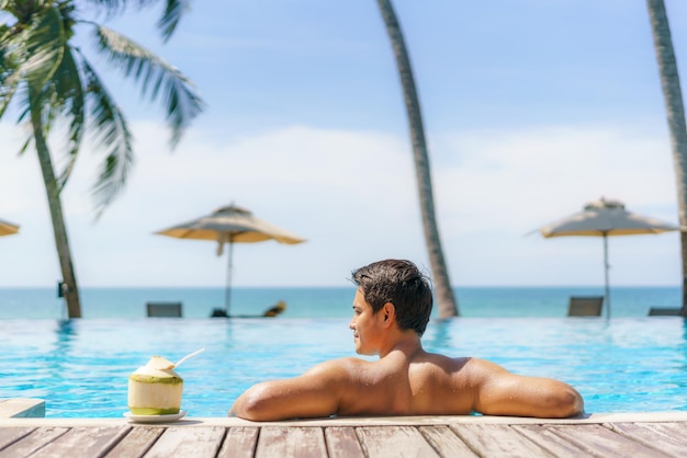 Koncepcja Wakacji Letnich Podróży, Podróżnik Azjatycki Mężczyzna Z Kokosem Relaksuje Się W Luksusowym Kurorcie Hotelowym Z Basenem Bez Krawędzi Z Plażą Morską I Tłem Palmy