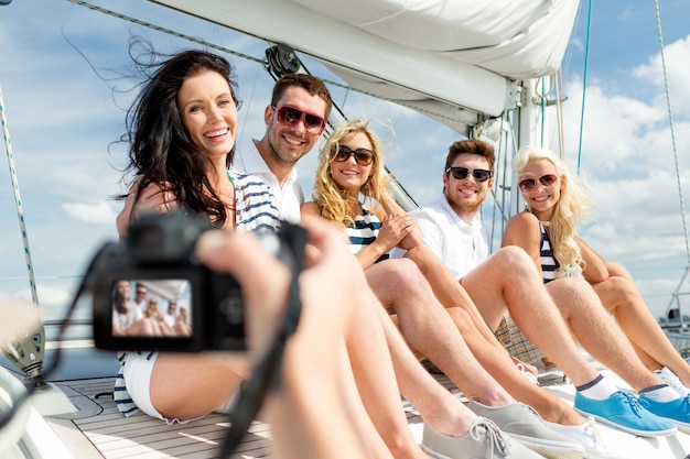 koncepcja wakacji, podróży, morza, przyjaźni i ludzi - uśmiechnięci przyjaciele siedzący na pokładzie jachtu i fotografujący