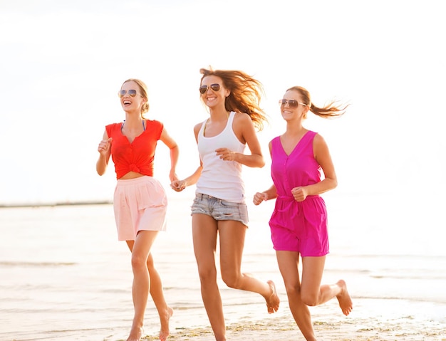 koncepcja wakacji, wakacji, podróży i ludzi - grupa uśmiechniętych młodych kobiet w okularach przeciwsłonecznych i codziennych ubraniach biegających po plaży