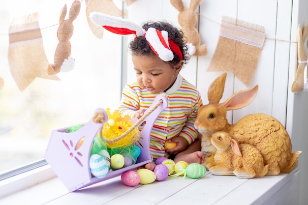 Koncepcja Wielkanocy afroamerykańskie małe dziecko w uszach zająca na głowie z jajkami i królikiem na oknie domu