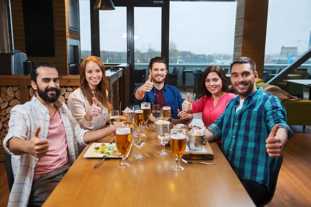 koncepcja wypoczynku, jedzenia, jedzenia i napojów, ludzi i wakacji - uśmiechnięci przyjaciele jedzący obiad i pijący piwo oraz pokazujący kciuki w restauracji lub pubie
