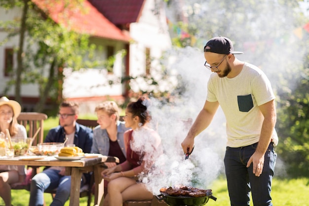 koncepcja wypoczynku, jedzenia, ludzi i wakacji - mężczyzna gotuje mięso na grillu dla swoich przyjaciół na letniej imprezie plenerowej