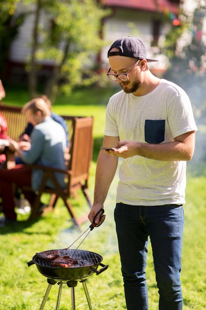 koncepcja wypoczynku, jedzenia, ludzi, technologii i wakacji - mężczyzna gotuje mięso na grillu i wysyła wiadomość tekstową na smartfonie na letniej imprezie plenerowej