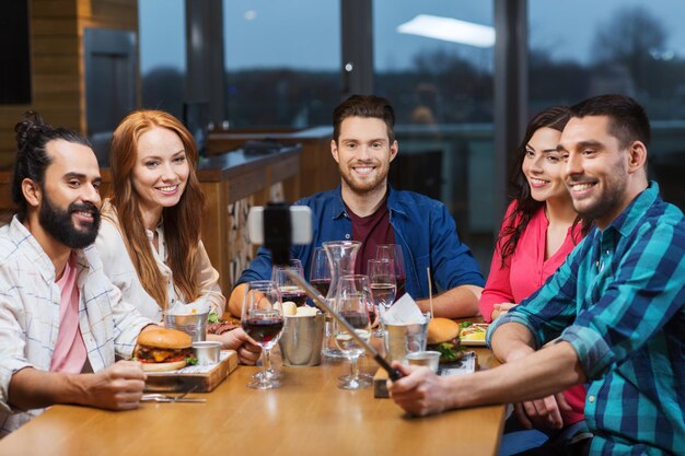 koncepcja wypoczynku, technologii, przyjaźni, ludzi i wakacji - szczęśliwi przyjaciele jedzą obiad i robią zdjęcia za pomocą smartfona selfie stick w restauracji