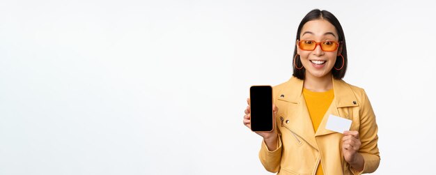 Koncepcja zakupów online i ludzi Stylowa azjatycka kobieta pokazująca ekran telefonu komórkowego i aplikację smartfona z kartą kredytową stojącą na białym tle Skopiuj miejsce