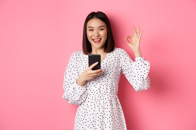 Koncepcja zakupów online i urody. Zadowolona azjatycka klientka Wyświetlono w porządku, dokonując zakupu w internecie na smartfonie, stojąc na różowym tle.