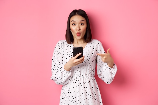 Koncepcja zakupów online i urody. Zdziwiona azjatycka kobieta wskazująca palcem na telefon komórkowy, polecająca aplikację na smartfona, stojąca na różowym tle.