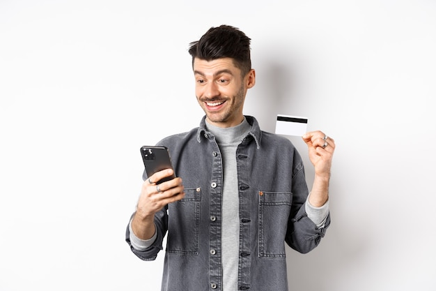 Koncepcja zakupów online. Uśmiechnięty facet płaci na telefon komórkowy, pokazując plastikową kartę kredytową, kupując w internecie, białe tło.