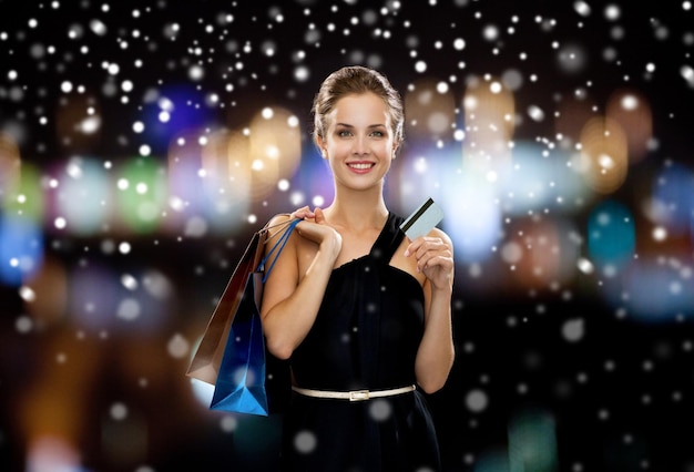koncepcja zakupów, sprzedaży, bankowości, pieniędzy i wakacji - uśmiechnięta kobieta w sukience z torbami na zakupy i kartą kredytową na czarnym tle