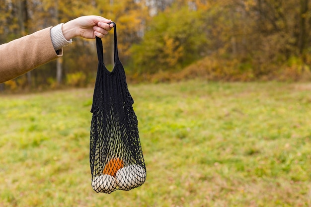 Zdjęcie koncepcja zakupów zero odpadów. bez plastiku. kobieca ręka trzyma czarną torbę eco string z pomarańczowymi i białymi dyniami na tle przyrody